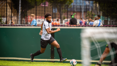 A male footballer is dribbling the ball and sprinting down the sideline. In the foreground you can see the goal with the keeper poised to save the ball.