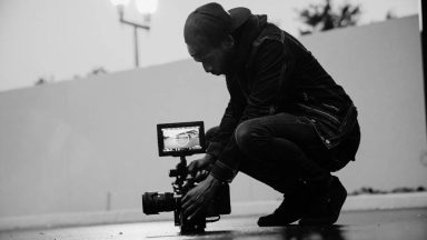 Black and white photo of a camera operator setting up a shot. They are in an outdoor carpark area, crouched down with the camera sitting on the ground.