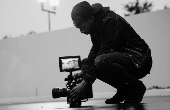 Black and white photo of a camera operator setting up a shot. They are in an outdoor carpark area, crouched down with the camera sitting on the ground.