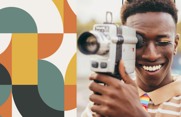A young Black filmmaker is holding a Super 8 camera up to his eye, whilst smiling.