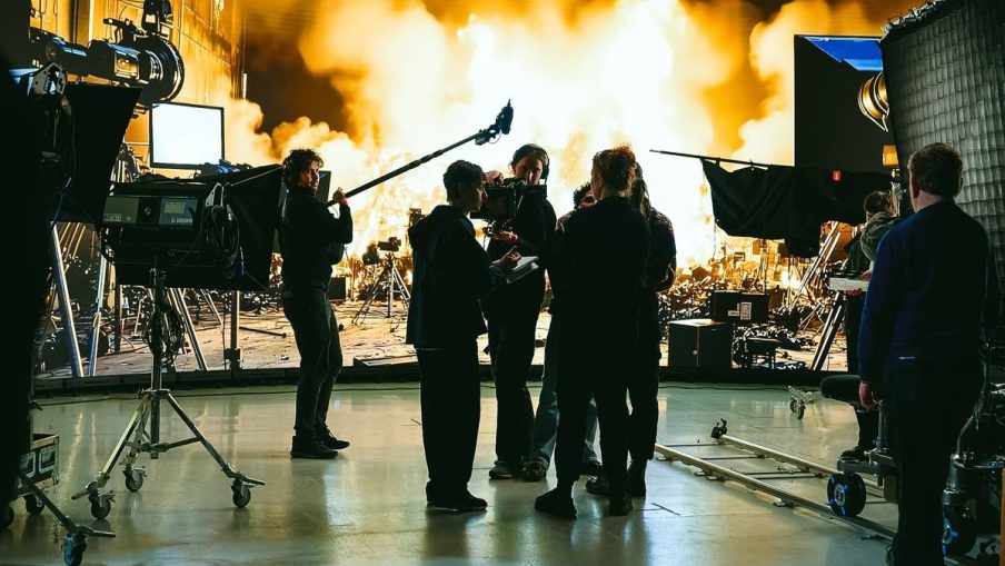 A film crew collaborates in a studio, with dramatic flames and smoke glowing in the background, surrounded by filming equipment.