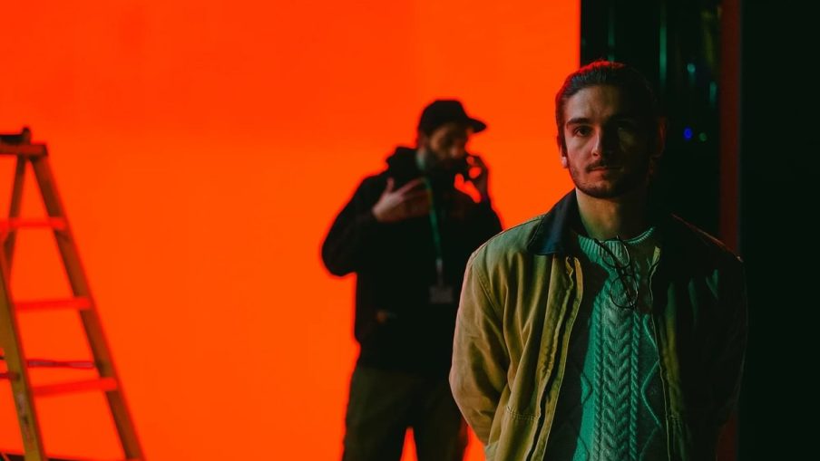 A person stands in front of a vibrant orange backdrop, while a figure in a black hoodie is partially visible behind them, holding a phone.