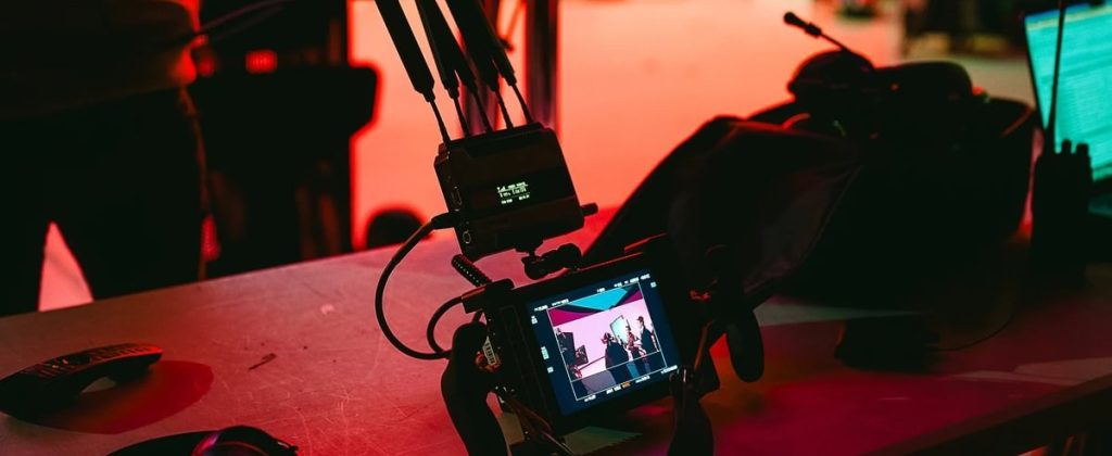 A video monitor displays a recording of a performance, surrounded by camera equipment and studio setup bathed in red light.