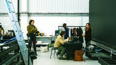 A busy studio space with people working at a table surrounded by clothing racks, a ladder, and various equipment scattered around.