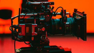 A close up of a film camera on a tripod in a brightly lit film studio.
