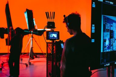A person monitors a camera setup in a studio with vibrant orange sound-stage background and lighting equipment, creating a dynamic filming environment.