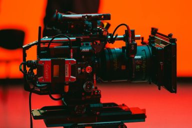 A close up of a film camera on a tripod in a brightly lit film studio.