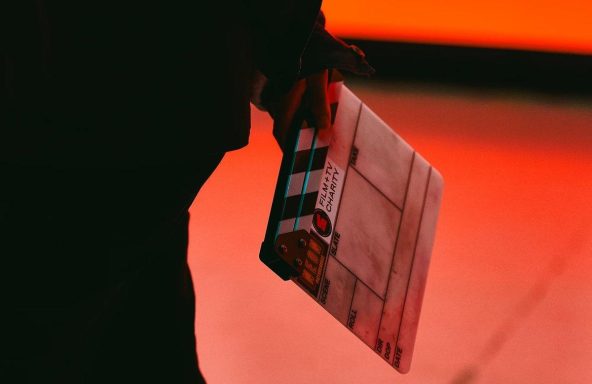 A close up of a person holding a clapperboard behind their back. On the board is the Film and TV Charity logo