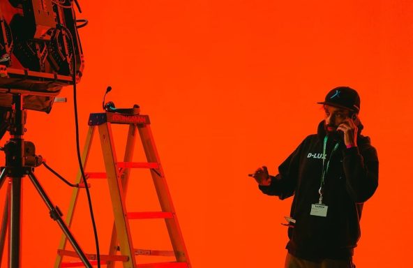 A crew member is on the phone in a brightly lit film studio. There is lighting equipment around him.