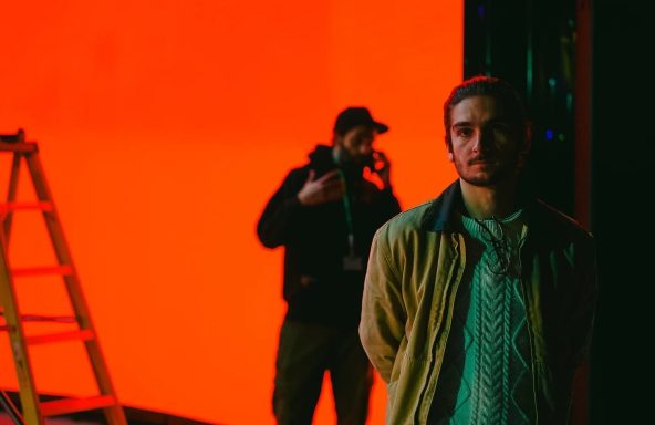 A person stands in front of a vibrant orange backdrop, while a figure in a black hoodie is partially visible behind them, holding a phone.