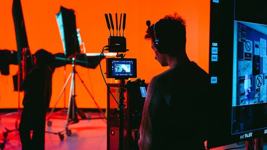 A person monitors a camera setup in a studio with vibrant orange sound-stage background and lighting equipment, creating a dynamic filming environment.