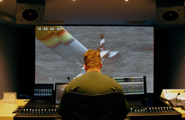 A TV editor is sat to an editing desk that has a large mixing desk. In front of him is a large playback screen with the image of a small catamaran sailing boat.