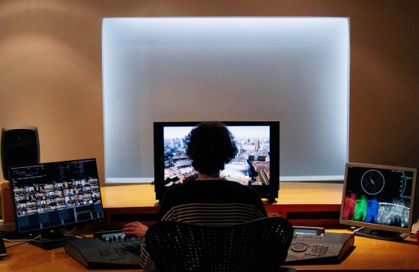 A colourist is sat in a dimly lit editing suite. They are silhouetted by the main playback monitor and they also have two screens to either side with various editing software screens open.