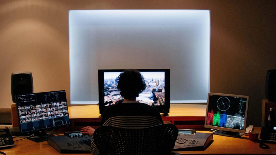 A colourist is sat in a dimly lit editing suite. They are silhouetted by the main playback monitor and they also have two screens to either side with various editing software screens open.