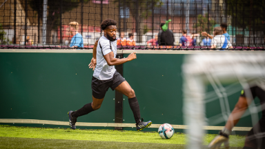 A male footballer is dribbling the ball and sprinting down the sideline. In the foreground you can see the goal with the keeper poised to save the ball.