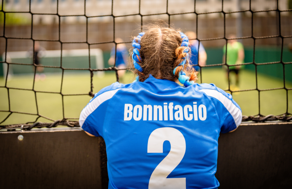 A football player is standing on the side-lines of a 5-a-side football match at the Broadcast Fives. They are wearing a blue sports top with the number 2 and the name Bonnifacio printed on the back.