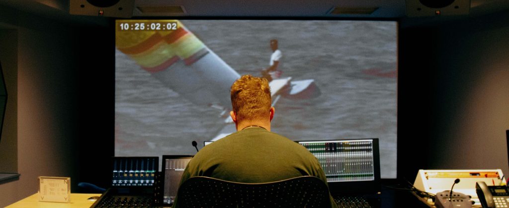 A TV editor is sat to an editing desk that has a large mixing desk. In front of him is a large playback screen with the image of a small catamaran sailing boat.