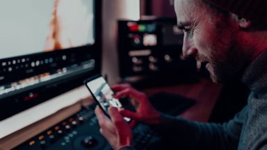 A man is sat to an editing desk in a suite. He is holding his phone and zooming in on a photo.