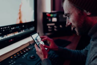 A man is sat to an editing desk in a suite. He is holding his phone and zooming in on a photo.