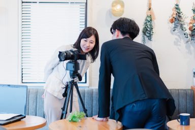 A videographer is setting up his camera on a tripod in the corner of a cafe. The client is leaning in to look at the camera monitor. There are dried flowers hanging on the white wall in the background, above a row of blue padding bench seating.