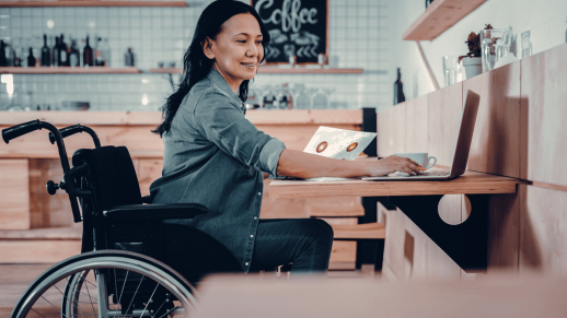 A worker is sat in a wheelchair to a desk. They have a laptop in front of them and they are holding a piece of paper with graphs printed on it.