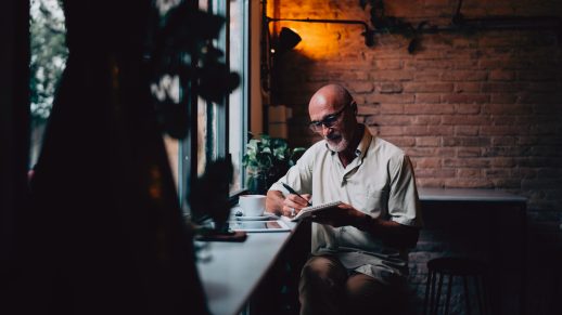 Casual dressed man wearing glasses is sat in a coffee shop. He is making notes in a pad that he is holding.