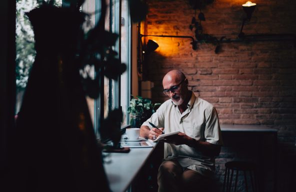 Casual dressed man wearing glasses is sat in a coffee shop. He is making notes in a pad that he is holding.