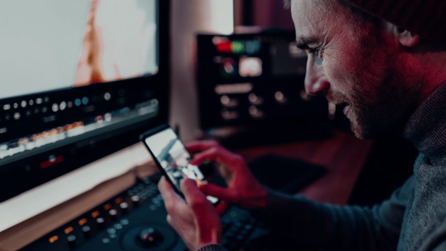 A man is sat to an editing desk in a suite. He is holding his phone and zooming in on a photo.