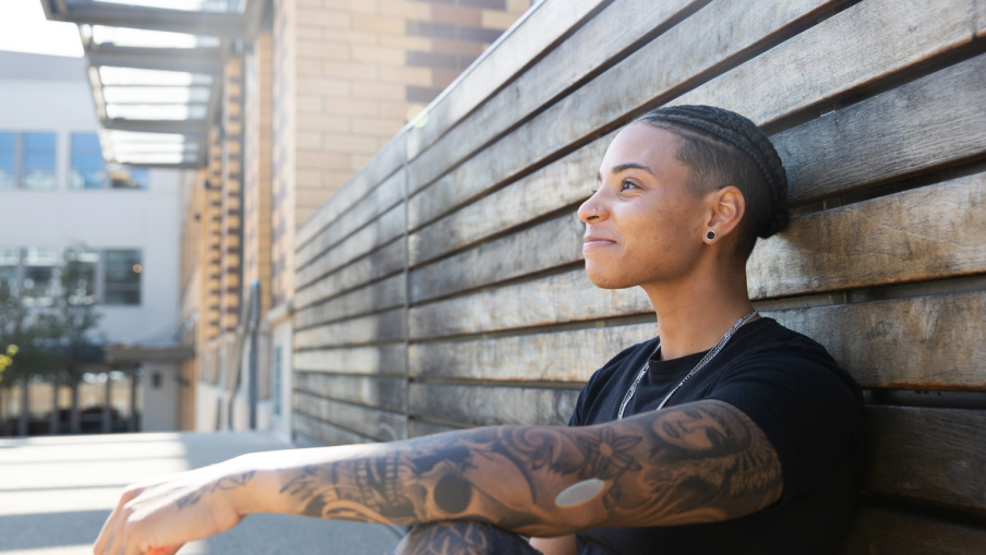 A young tattooed person is sitting on the floor with their back against a wooden slatted fence. They are looking out into the open and smiling.