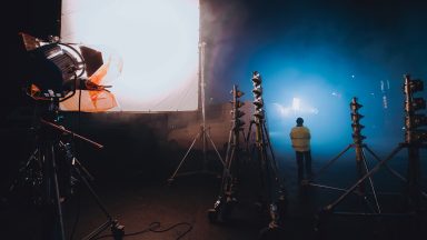 Lighting equipment illuminates an outdoor set with a glowing blue light. In the background is a crew member facing away, they are wearing a high-viz jacket.