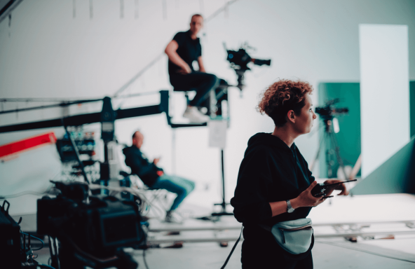 A small crew are set up in a bright, white studio. In the foreground, a director is standing whilst holding a walkie-talkie. In the background there is a camera operator sitting at the end of a camera crane awaiting direction.