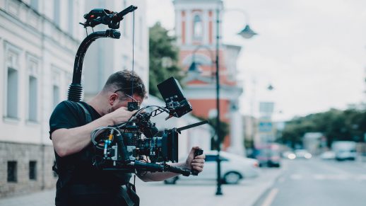 Director of photography with a camera connected to a back-brace, in his hands is the camera. He is looking through the viewfinder. In the background is an empty street.