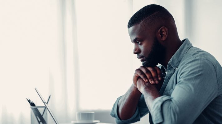 Thoughtful writer is sat at a desk with his head resting on his hands. He has a laptop in front of him with a notepad to the side.