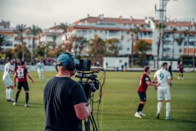 A camera operator is standing behind a camera set up for an outside broadcast on the side-lines of a football pitch. They are tracking football players that are on the pitch mid-game.