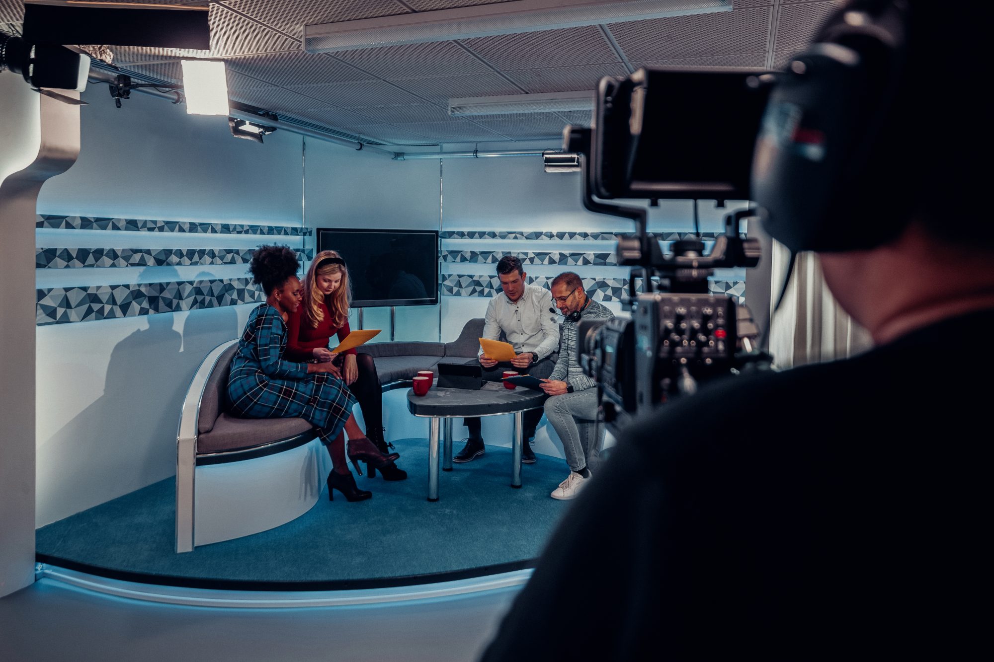 Over the shoulder view of a TV show in the process of being filmed in a studio. The presenters are sitting on the studio sofa while looking at notes and talking to the producer, while the camera man is preparing to start filming again.
