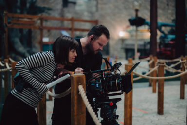 Female director at work on the set. She is leaning over to check a shot that the camera operator has set up. In the background you can see other behind the scenes equipment.
