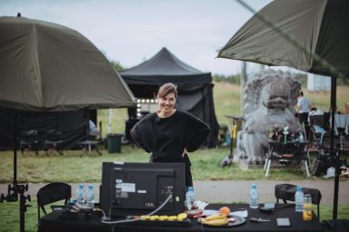 A female director standing and reviewing a playback monitor whilst filming a movie. She is looking up and smiling at the camera. In the background there are other behind-the-scenes tents and other equipment and props.