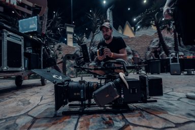 A focus puller on set, he is crouching down and testing the equipment. In front of him is a film camera that is resting on the floor.