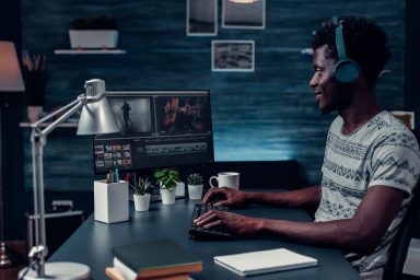 Young make video editor is sat to their desk at home editing on a computer. He is wearing headphones and a white, striped t-shirt.