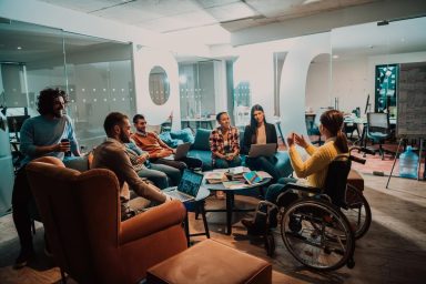 A group of people are sat in an modern and bright office meeting space. One woman in a wheelchair is sat at the front, addressing the others.
