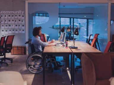 A man working at a desk in wheelchair and using a desktop computer in an open space modern office.