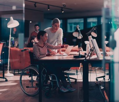 Two women at a desk, one is in a wheelchair and the other standing by their side. They are both looking at the laptop screen.