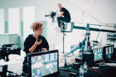 Production Manager is holding a walkie talkie. She is looking at the crew. In the background, there is a jib-mounted camera operator.