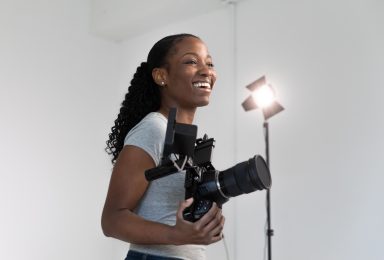 A female photographer is standing in a bright, white studio space. She is smiling and holding a DSLR camera with a field monitor attached to the hot shoe. Behind her is a studio light that is shining towards the camera.