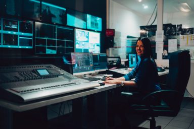 Producer working in a broadcast control room. She is sat at a large desk, full of equipment and she is facing the camera and smiling.