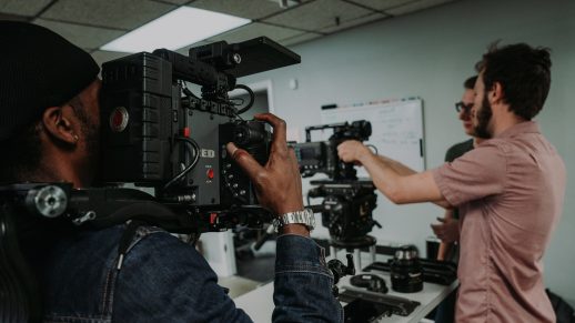 Close up of a camera operator holding a RED camera. The camera is pointing at two other crew members who are prepping an additional camera.