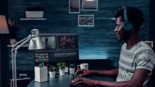 Young make video editor is sat to their desk at home editing on a computer. He is wearing headphones and a white, striped t-shirt.