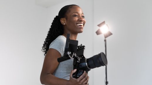 A female photographer is standing in a bright, white studio space. She is smiling and holding a DSLR camera with a field monitor attached to the hot shoe. Behind her is a studio light that is shining towards the camera.