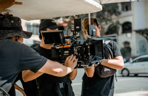 A camera operator is walking down a street whilst holding a large RED camera rig. To either side of them is an assistant that is supporting the camera whilst another follows behind holding a reflector board above the camera.