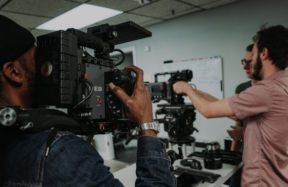 Close up of a camera operator holding a RED camera. The camera is pointing at two other crew members who are prepping an additional camera.